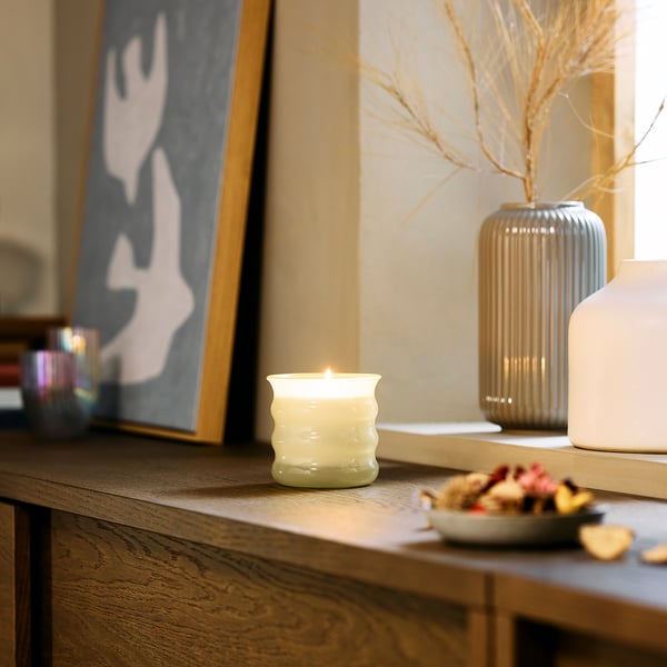 Cosy room decor: lit JUVELTRAST vanilla candle in green glass sits on wood shelf, near vases and fruit. Small, warm, calming.