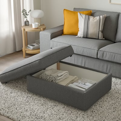 Modern living room: grey sectional sofa with yellow pillow, wooden side table with lamp and books. Ottoman open, showing blanket and magazines inside.