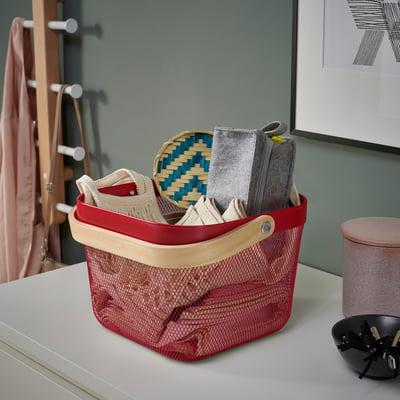 Red mesh basket with wooden handle on counter. Used for storing and organising items, like a grey felt container and woven baskets.