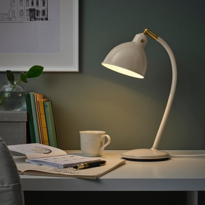 White RÖDFLIK desk lamp with brass on workspace. Includes mug, notebook, pen, books, and wall picture.