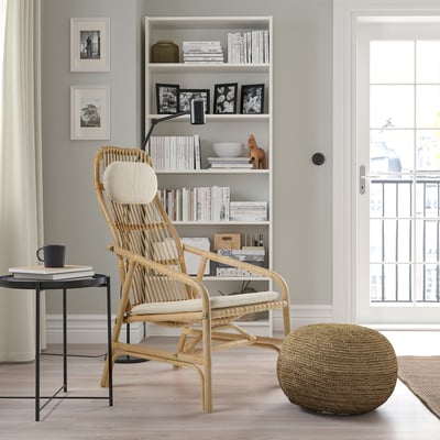Natural rattan chair, side table with books and mug, bookshelf, and window with curtains.