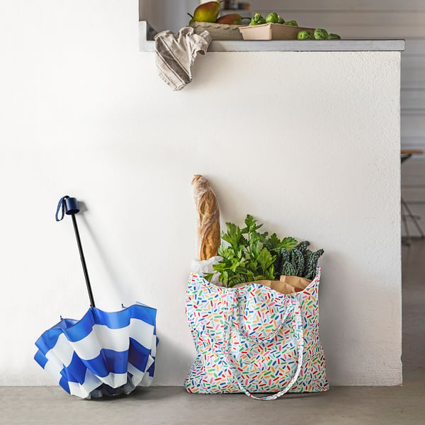 A colourful reusable shopping bag filled with groceries, leaning against a white cabinet. Beside it, a folded blue and white bag with a blue handle.