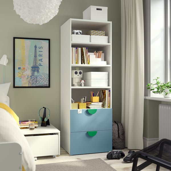 SMÅSTAD bookcase in light blue with green accents, three shelves, two drawers, near paris-themed artwork, bed, and black chair.