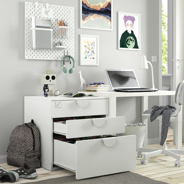 Childs desk with open drawers, laptop, headphones, backpack. Pegboard stores supplies. Wall has artwork.