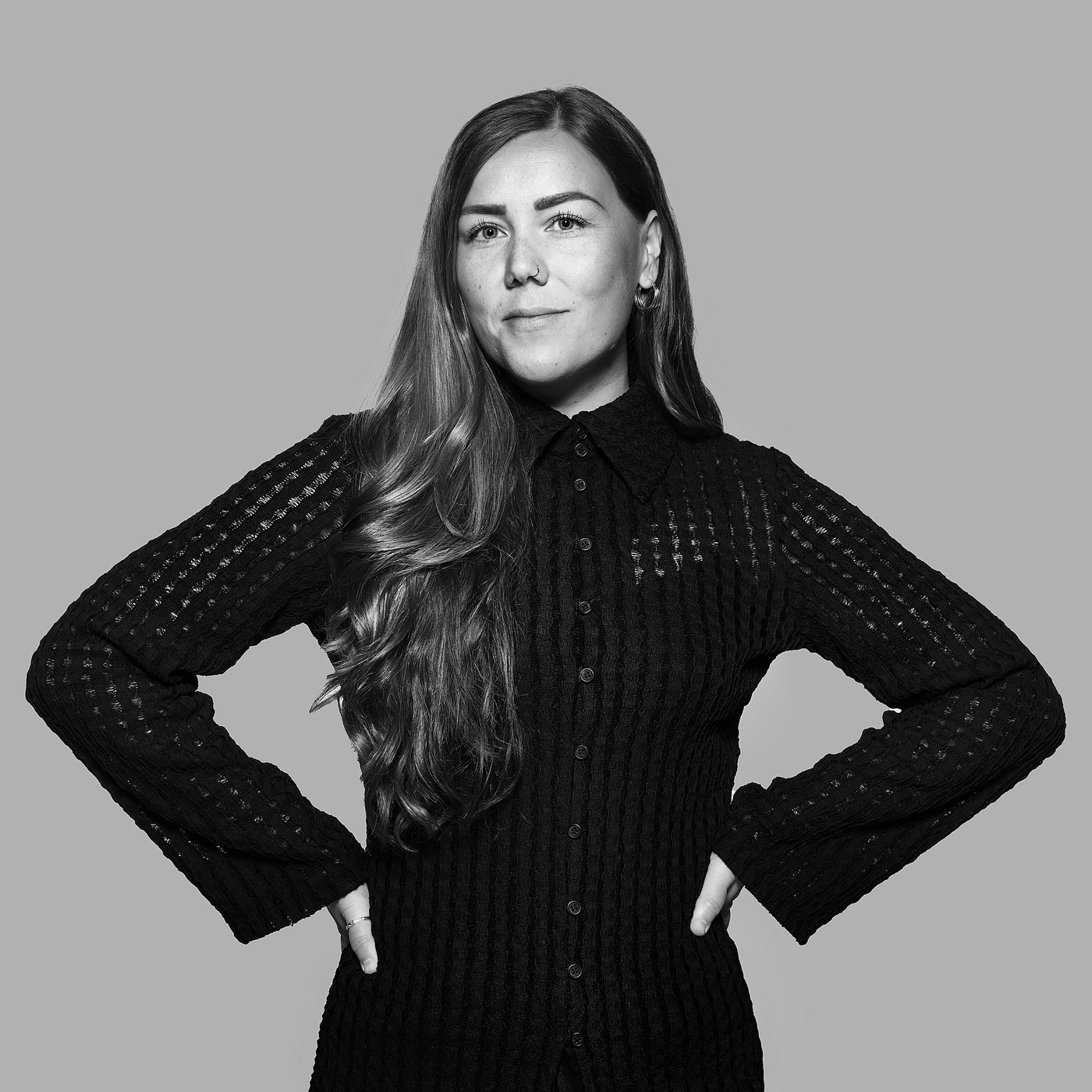 Black-and-white portrait of the designer with long hair, confidently posing with hands on hips, wearing a dark sweater.