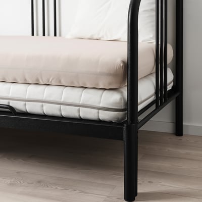 Black metal bed with beige mattress and pillows on wooden floor.