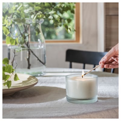 Hand lights scented candle in glass jar on table. Jars hold white candle, green plant in background.