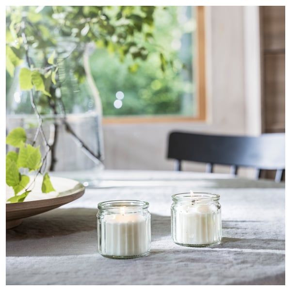 Two lit glass candles on a table, with a window and plant in the background. The candles are white and the glass has a ribbed design.