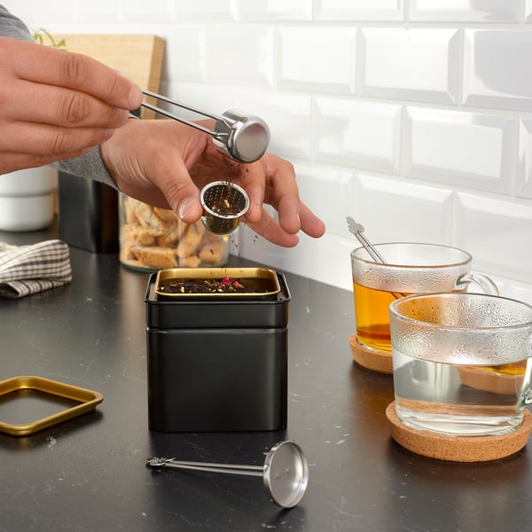 Person using ÄNGSBLÅVINGE tea infuser, pouring tea into mug from black tin container, showcasing its functional design.