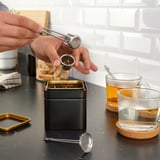 Person using ÄNGSBLÅVINGE tea infuser, pouring tea into mug from black tin container, showcasing its functional design.