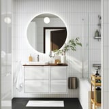 Modern white bathroom with round mirror, sleek vanity, and glass shower. White towels and plants decorate.