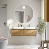 Modern bathroom with large round mirror, dual wooden vanity, white basins, and tall houseplant.