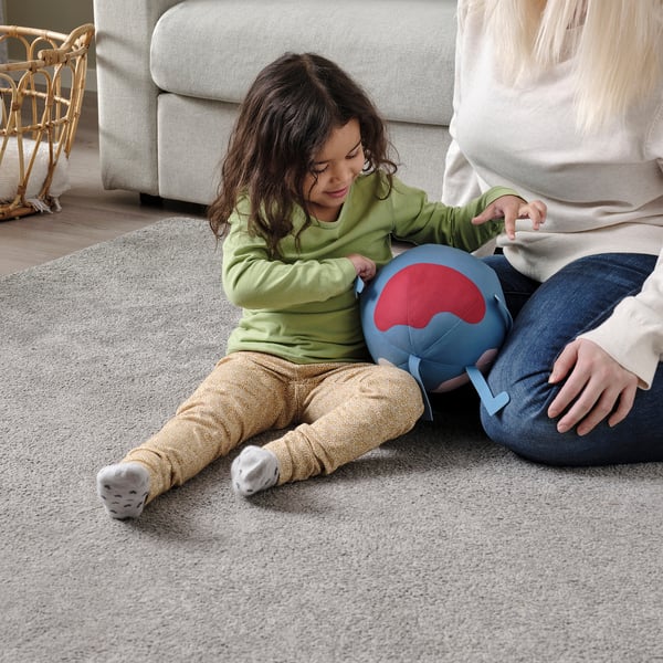 Child playing with soft earth toy on floor. Adult beside. Light gray carpet. Light-colored couch. Wicker chair visible.