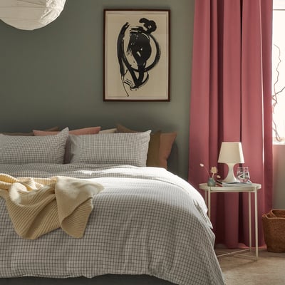 Bedroom with green walls, large ÅKERFIBBLA white & black checkered duvet set, red curtains, side table, lamp, basket & art above bed.