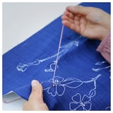 Person hand-embroidering blue cushion cover with white floral design. Thread is visible as they work on intricate stitching.