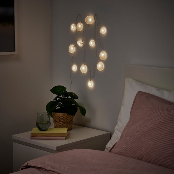 Cozy bedroom with soft AKTERPORT lights, peach pillows, plant, books, and nightstand.