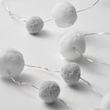 Image shows a close-up of decorative string lights featuring several round, fuzzy pom-pom bulbs connected by thin wires.