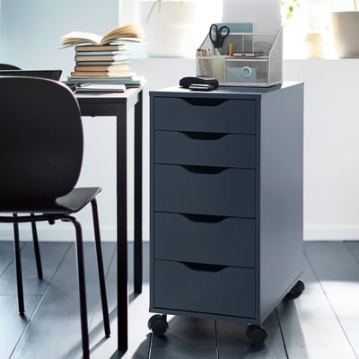 Gray ALEX drawer unit with five drawers on castors near a black chair and desk, storing office supplies.