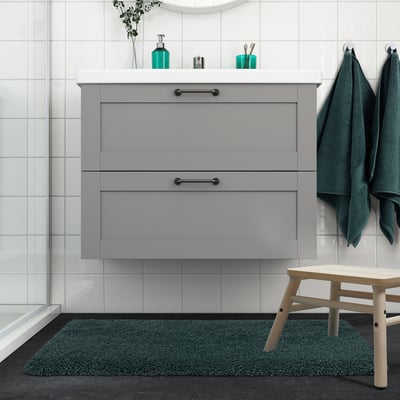 Modern bathroom with ALMTJÄRN grey-turquoise bath mat, wooden stool, grey cabinet, and hanging towels.