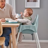 A baby sits in an ANTILOP highchair, drinking from a cup while a parent nearby assists at a wooden table.