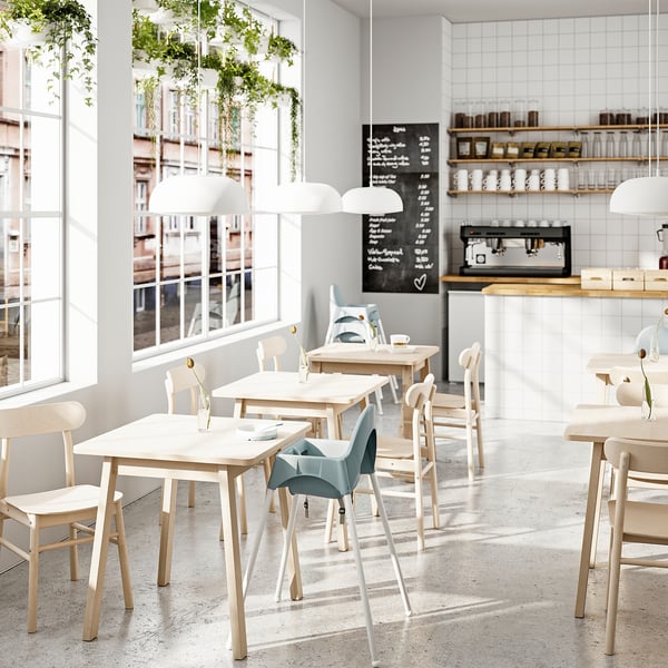 Modern café with light wood tables, chairs, large windows, hanging plants, and a counter with shelves and a coffee machine. Bright, airy atmosphere.