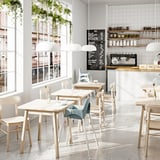 Modern café with light wood tables, chairs, large windows, hanging plants, and a counter with shelves and a coffee machine. Bright, airy atmosphere.