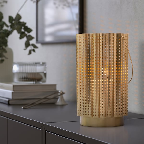 Gold, decorative lantern with handle on a table. Illuminates with warm light through punched holes, part of AROMATISK series.