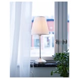 A lamp with a beige shade on a window sill, diffused light, white curtains, and a green plant.