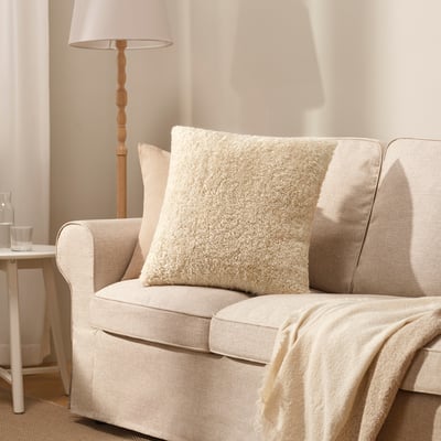Cozy beige ÅSJORDFLY square throw pillow on light sofa, soft teddy fur texture.