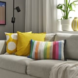 Bright yellow and multicolored striped cushions on a grey couch, with a potted plant beside, near a window with curtains.