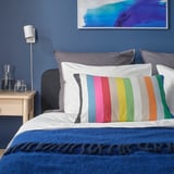 Bed with BÄCKMÄRKEN multicolor striped cushion, blue wall, abstract frame.