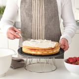 A person ices a cake on a rotating marble BAKGLAD stand, ensuring even coverage.