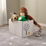 A white storage box with a tree print holds stuffed animals, and a baby bear doll is on the floor beside it.