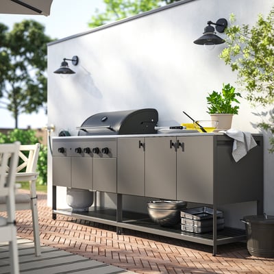 Modern outdoor kitchen with BÅTSKÄR grill, side burner, and storage. Hanging lights, plant, and seating nearby.