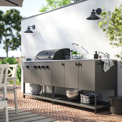 Outdoor kitchen setup with stainless steel grill, sink, and cabinets. Wall-mounted lights, plants, and brick patio.