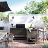 Modern outdoor kitchen with black units, gas grill, and seating. Lush greenery.