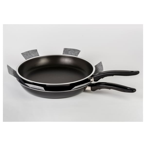 Stacked black frying pans with protective gray fabric strips, displayed on a light surface.