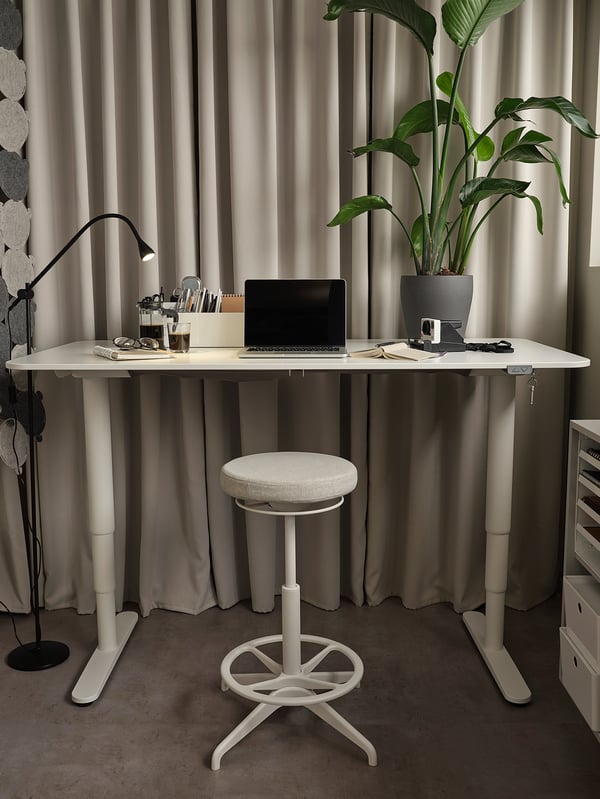A sleek white BEKANT desk with adjustable height, featuring a laptop, plant, and modern office accessories, complemented by a matching stool.