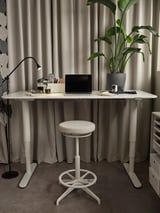 A sleek white BEKANT desk with adjustable height, featuring a laptop, plant, and modern office accessories, complemented by a matching stool.