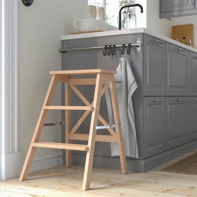 Wooden BEKVÄM step ladder against gray kitchen island. Foldable, light beech with three steps.