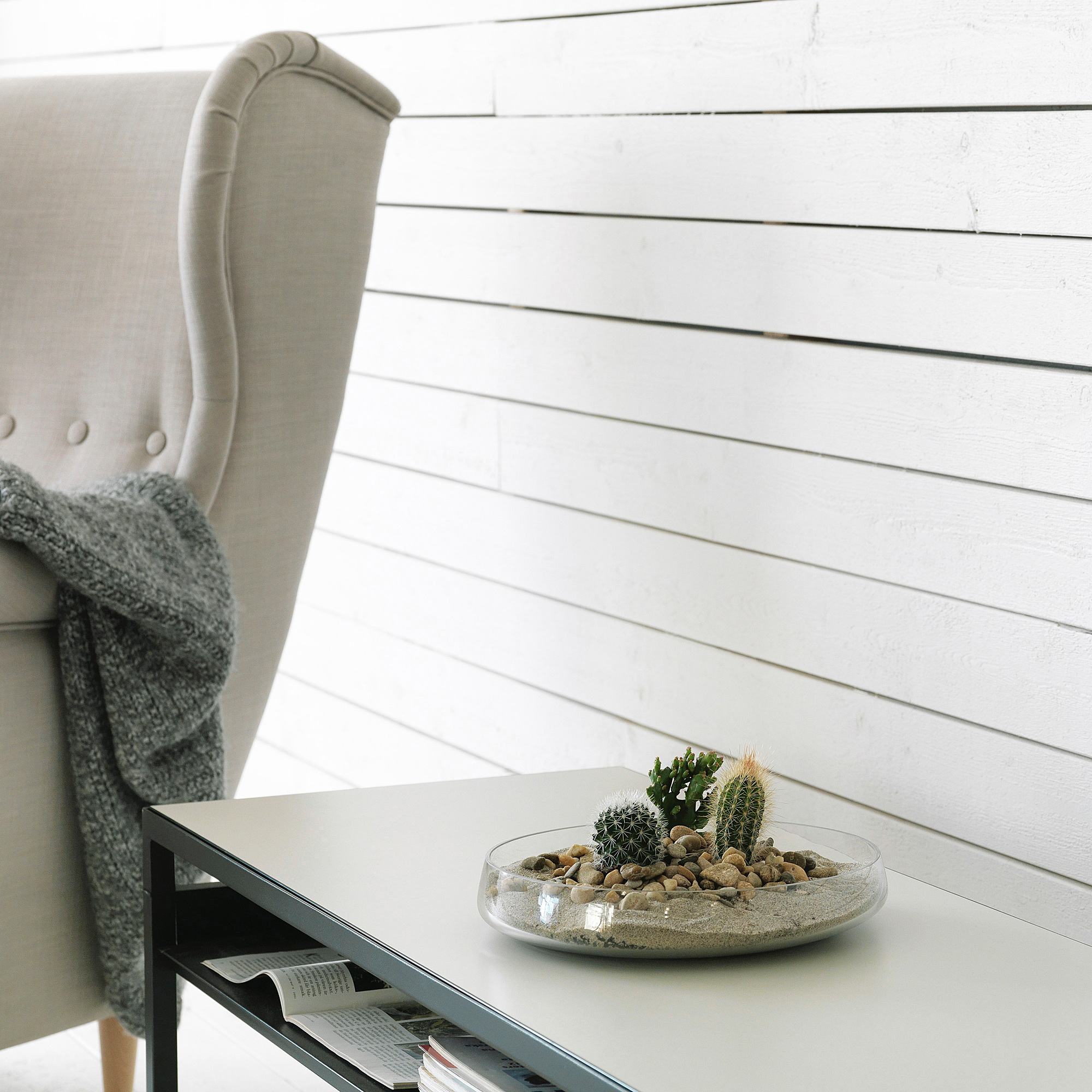 Modern chair with beige fabric and gray blanket, glass bowl with small plants and stones on white table against white wooden wall.