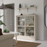 BERGSHULT beige shelf with glass doors, holding items, under wall shelf, near sofa and plant table.