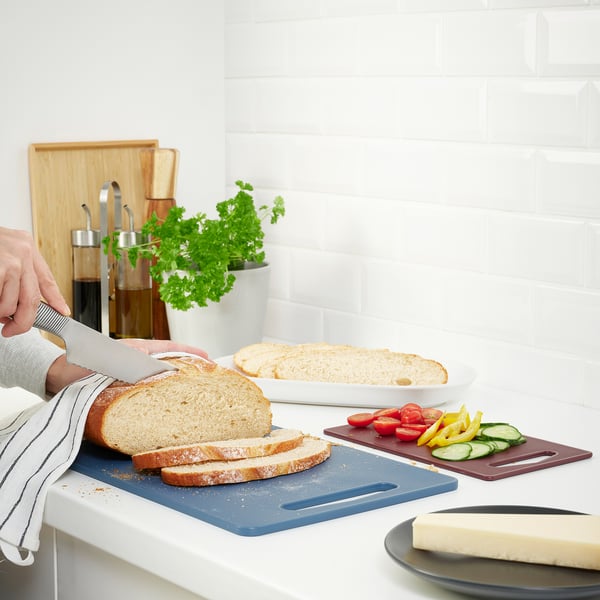 Person cutting bread on blue board with veggies and cheese nearby, white backsplash.