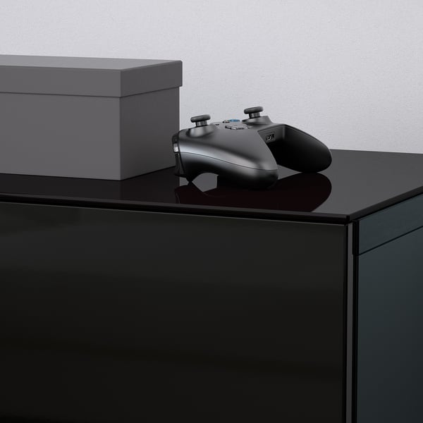 Black gaming controller resting on sleek, glossy glass-topped furniture, beside grey box.