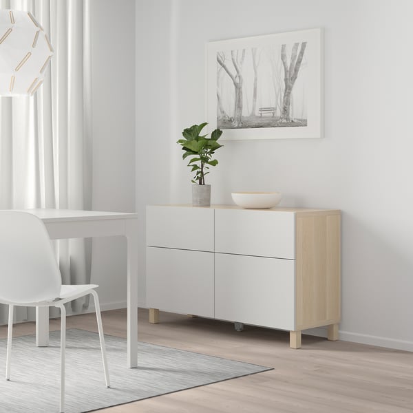 White BESTÅ sideboard with wooden legs, minimalist room, chair, desk, striped rug, black-and-white forest picture, plant, bowl. Spacious & elegant.