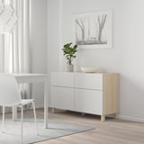 White BESTÅ sideboard with wooden legs, minimalist room, chair, desk, striped rug, black-and-white forest picture, plant, bowl. Spacious & elegant.