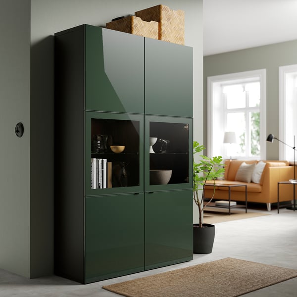 Large green BESTÅ cabinet with glass doors, holding bowls and books, beside an orange couch and window.