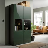 Large green BESTÅ cabinet with glass doors, holding bowls and books, beside an orange couch and window.
