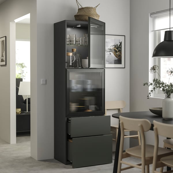 Black BESTÅ cabinet with glass doors showing drinkware and bowls, topped with a basket. Nearby dining table and chairs visible.