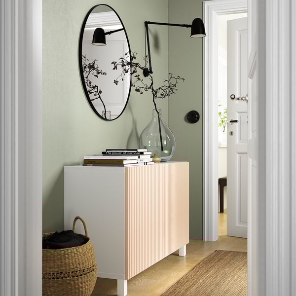 BESTÅ sideboard with white door, decorative items, green wall, mirror, lamps, basket, rug, doorway.